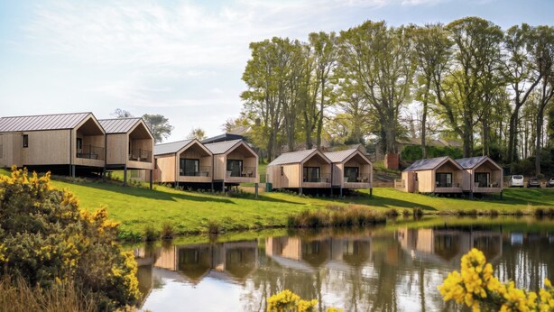 Une rangée de chalets en bois dotés de balcons, situés sur une pente herbeuse au bord d’un étang paisible, dont les eaux reflètent les chalets et les arbres environnants.