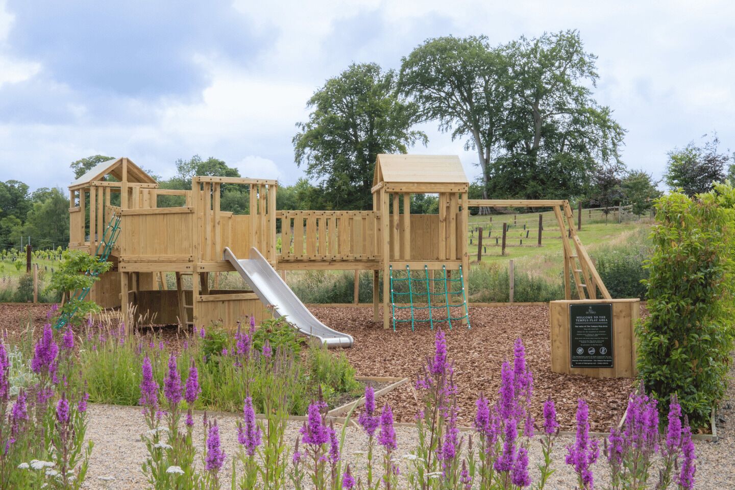 A wooden playground with a slide and climbing structures, surrounded by purple flowers and trees.