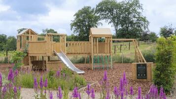 A wooden playground with a slide and climbing structures, surrounded by purple flowers and trees.