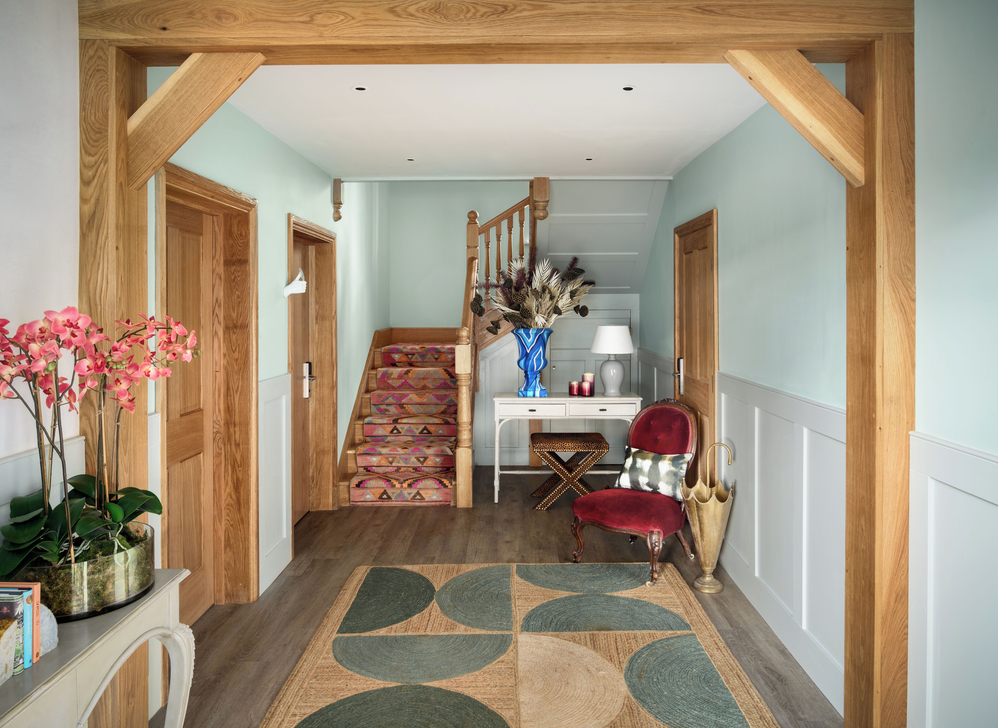 Un couloir avec un escalier en bois, un bureau avec une lampe, une chaise et un vase décoratif.