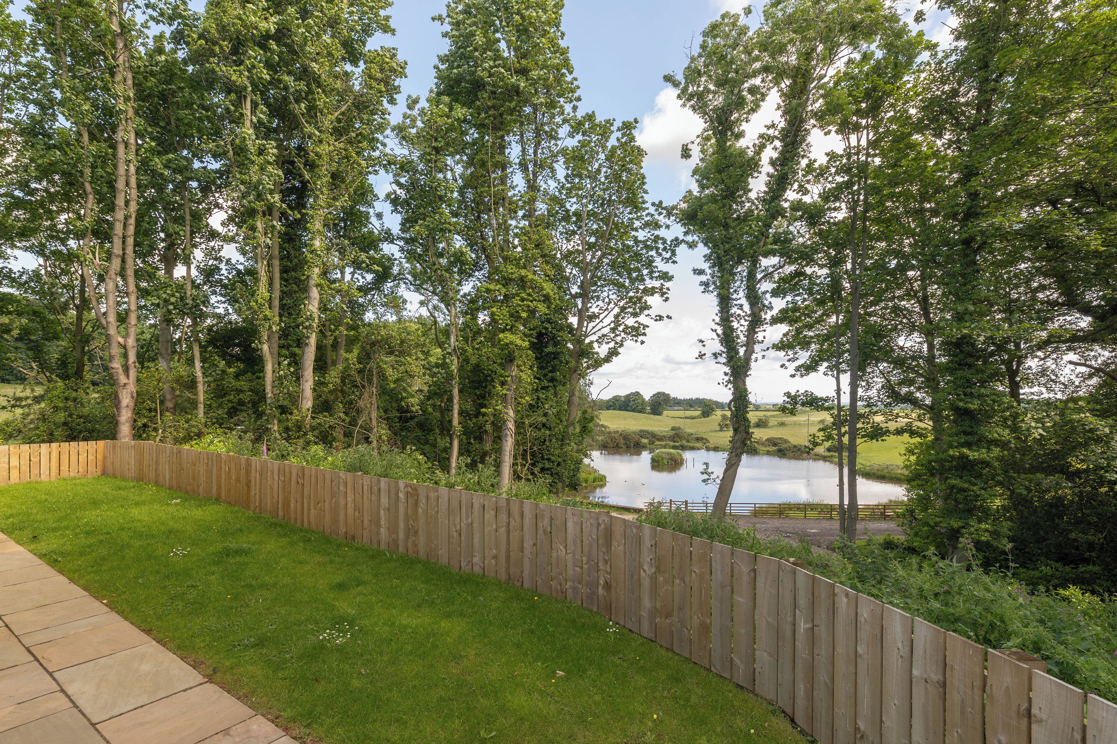 Een omheinde achtertuin met een gazon, bomen en een waterpartij op de achtergrond.