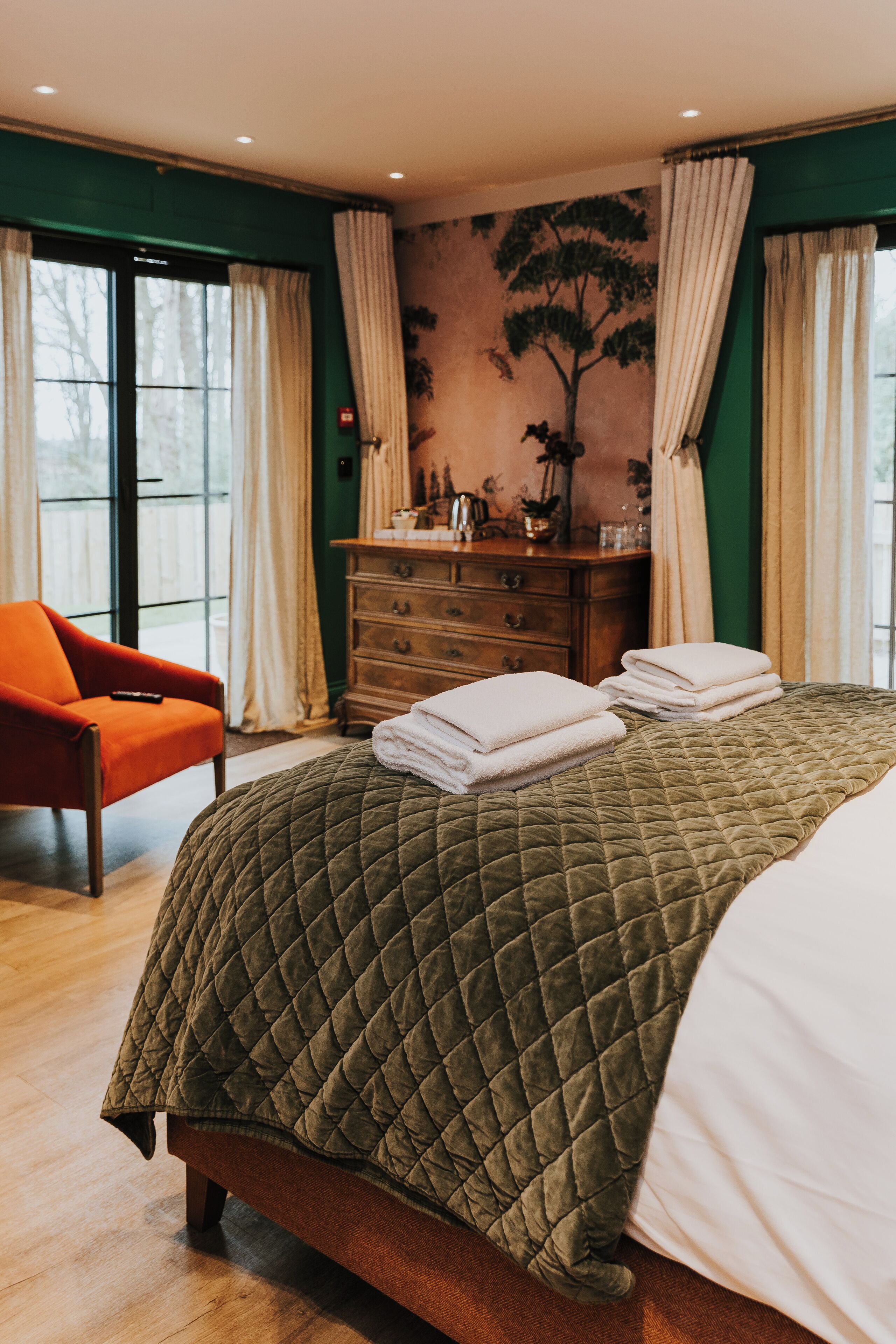 A hotel room with a large bed, a wooden dresser, a red armchair, and a view of the outdoors through a sliding door.