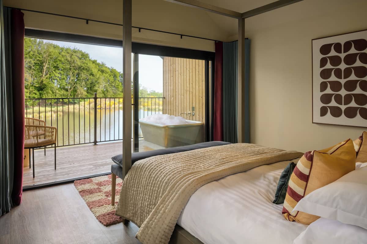 Un dormitorio con una cama grande, vistas a un lago, un balcón con una silla y una bañera.