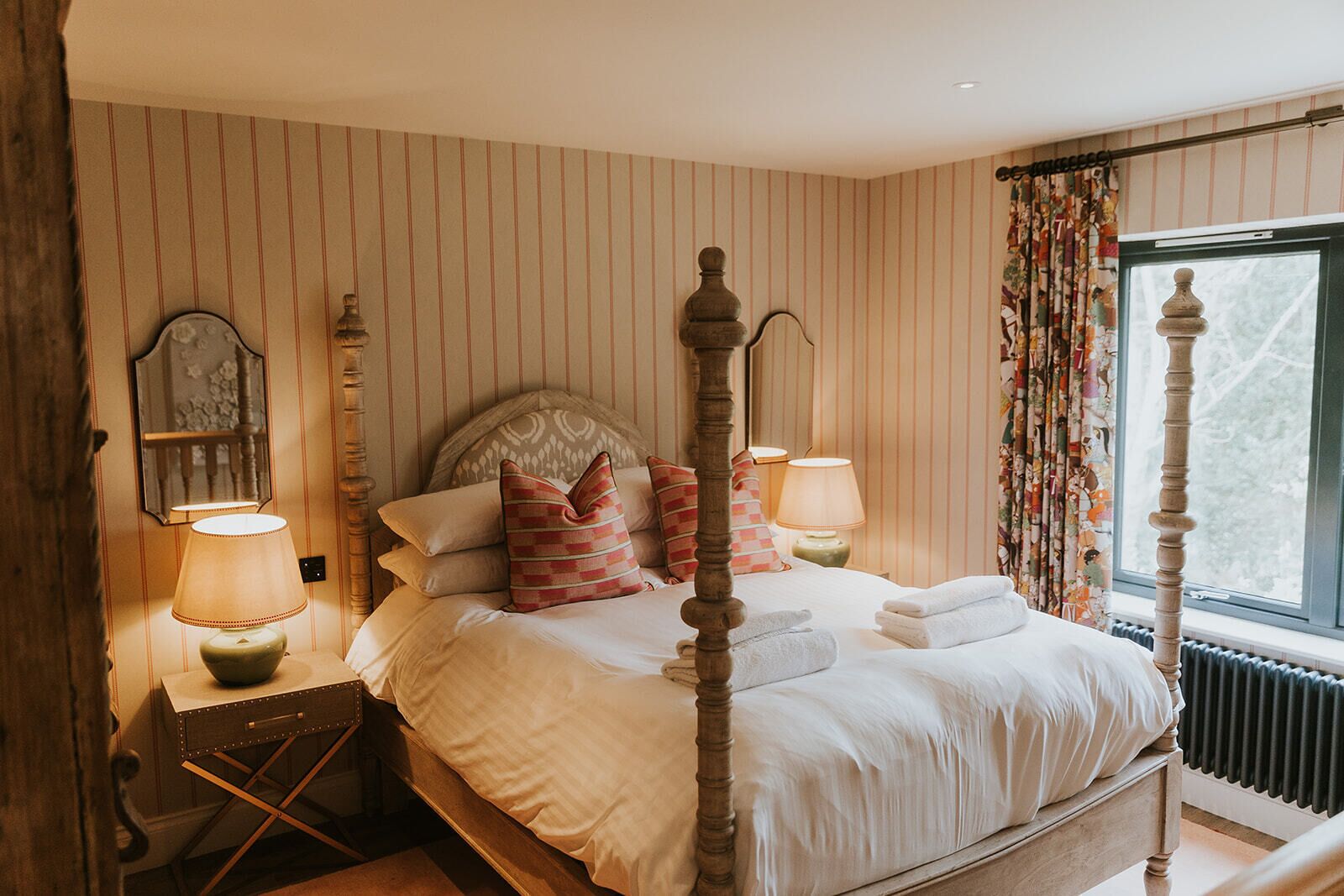 Un lit à baldaquin avec une tête de lit en bois, deux tables de chevet avec des lampes, un papier peint à rayures et une fenêtre avec des rideaux.