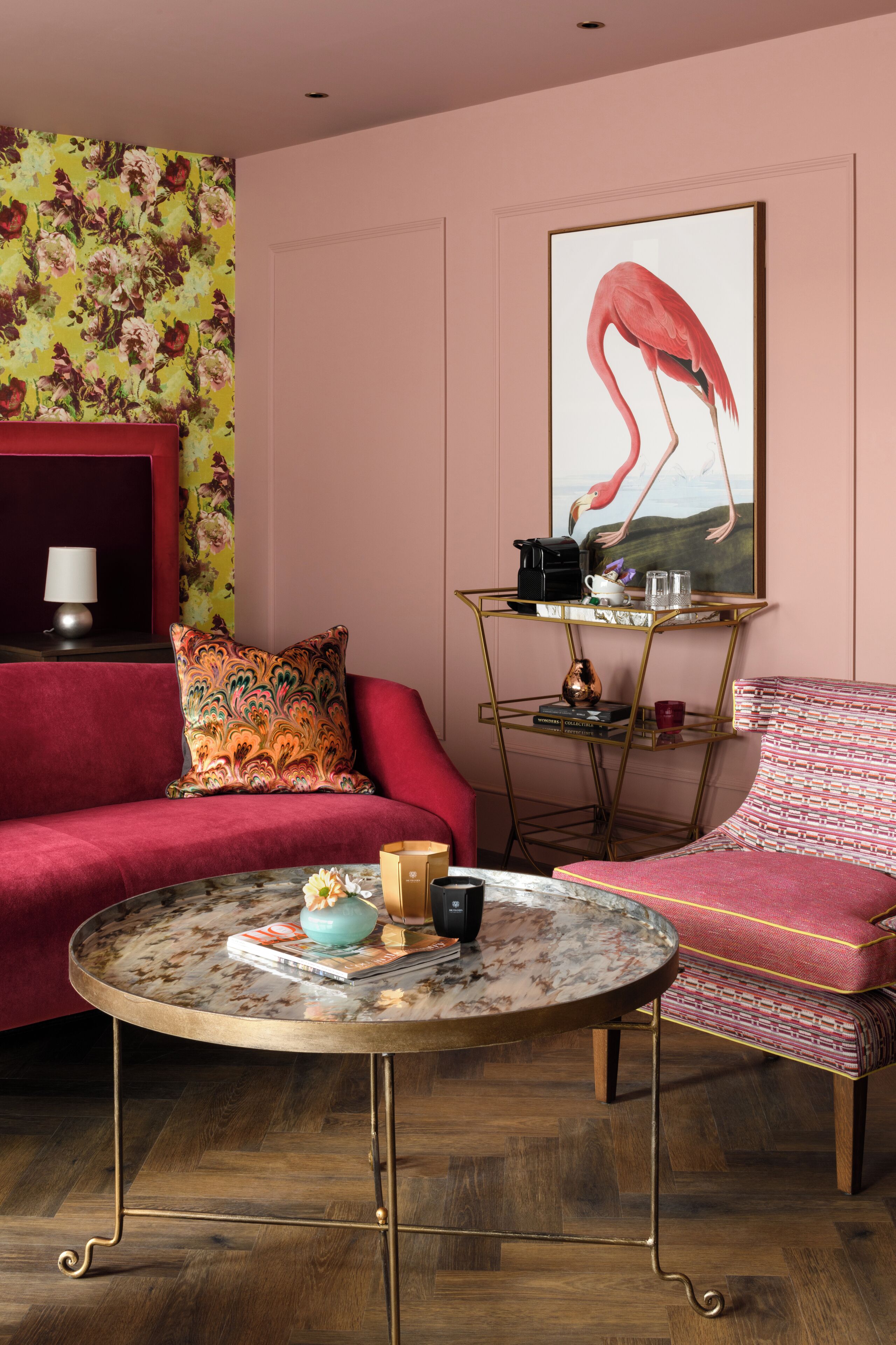 A living room with a red sofa, a patterned armchair, a round coffee table, and a wall with a floral wallpaper and a painting of a flamingo.