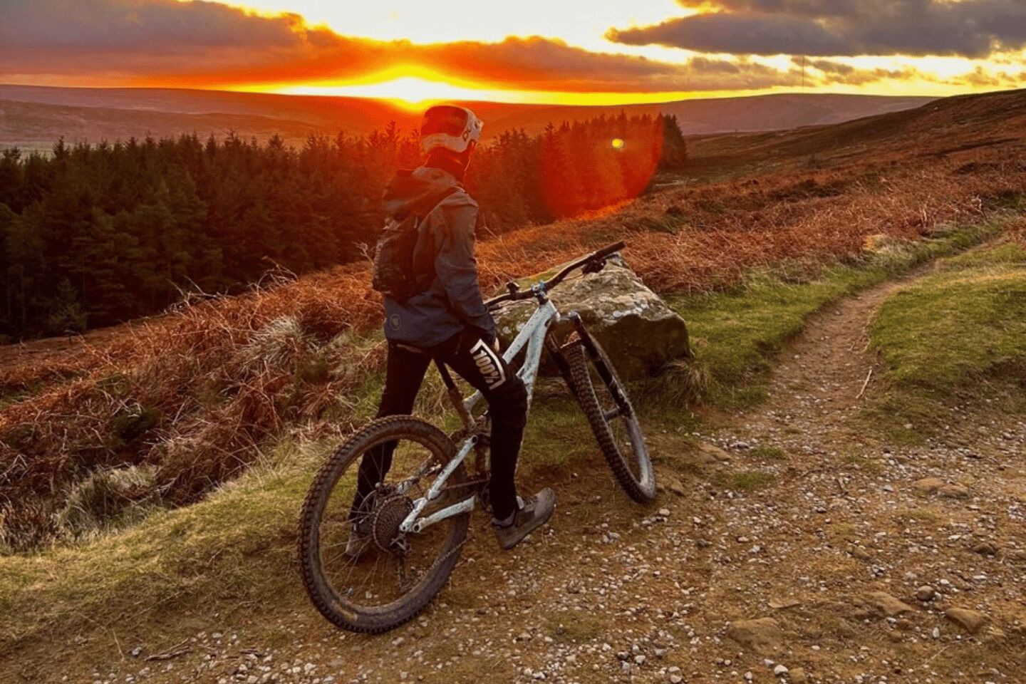 Een persoon staat naast een mountainbike op een onverhard pad, met een zonsondergang op de achtergrond.