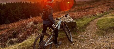 Een persoon staat naast een mountainbike op een onverhard pad, met een zonsondergang op de achtergrond.