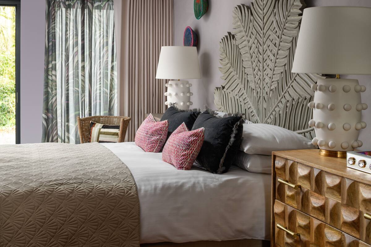 Un dormitorio con cama, dos lámparas de noche, una pared decorada con hojas, una silla de mimbre y una mesita de noche con un radio.