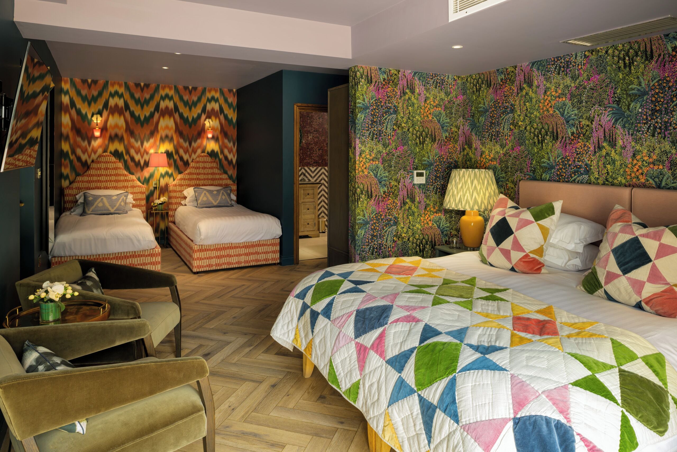 A hotel room with two beds, a sofa, a patterned wall, and a large window.