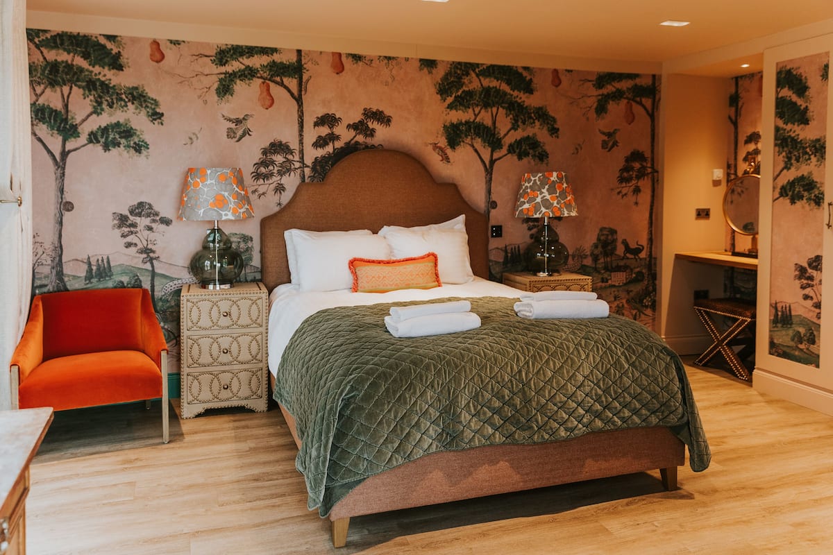 Un dormitorio con una cama grande, dos mesitas de noche con lámparas, una silla roja y un mural en la pared.