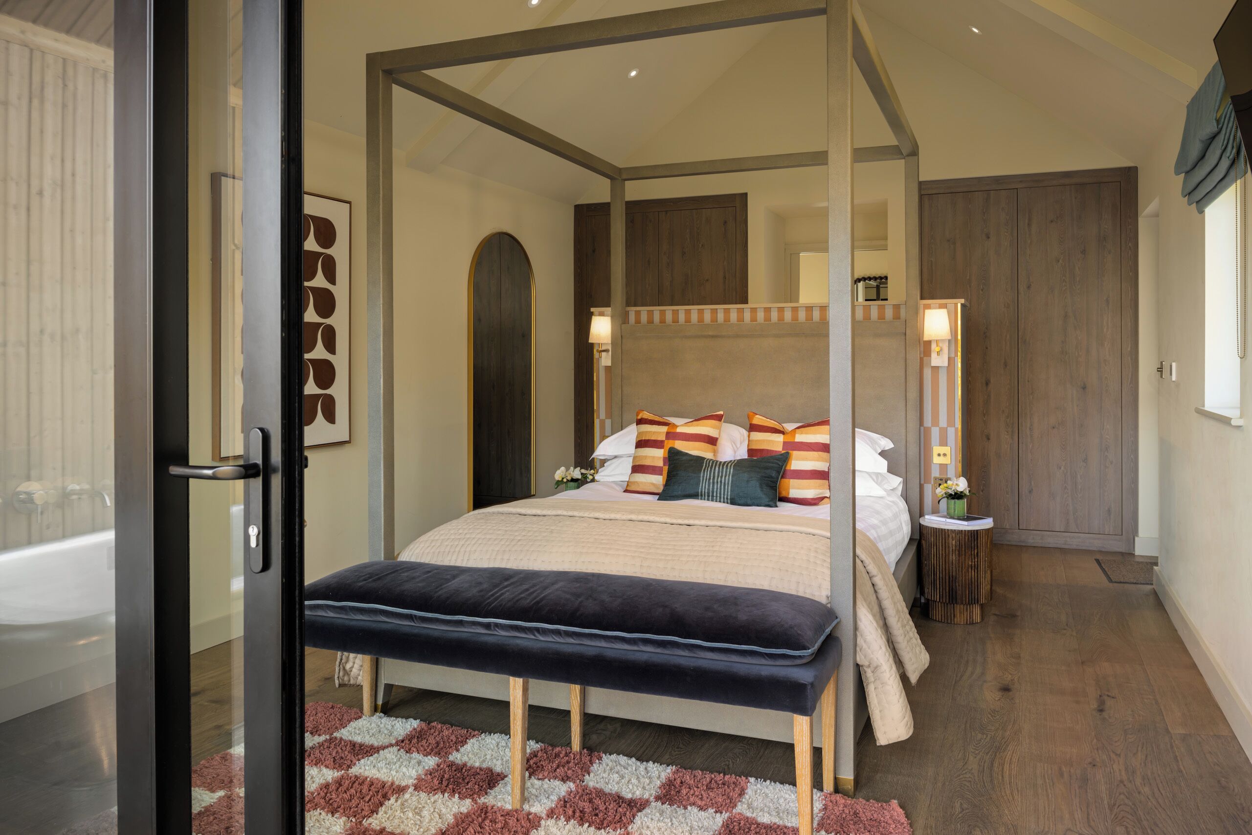 A modern bedroom with a four-poster bed, a bench, a small bedside table, and a view of a bathroom.