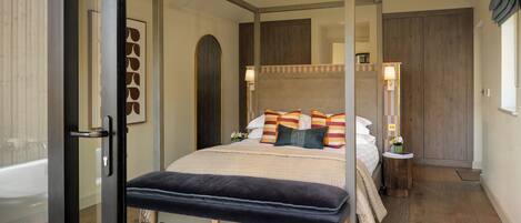 A modern bedroom with a four-poster bed, a bench, a small bedside table, and a view of a bathroom.