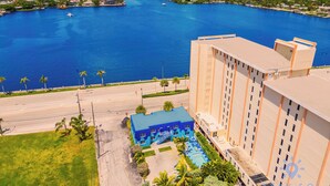 A high-rise hotel with a blue-roofed pool area, overlooking a large body of water with boats.