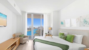 Moderni hotellihuone, jossa on suuri sänky, työpöytä, tuoli ja suuret ikkunat, joista avautuu näkymä kaupunkiin ja vesistöön.