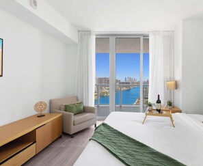 A hotel room with a bed, a small table with a wine bottle and glass, a chair, a sofa, a window with a view of a body of water and buildings, and a wall-mounted artwork.