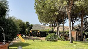 A well-maintained garden with a swing set, stone pathway, and mature trees. - Villa in Chia 5 minutes from the most beautiful beaches (Chia)