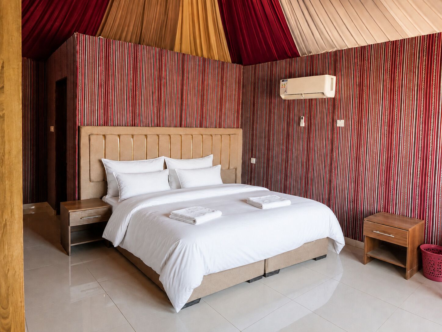 A bedroom with a large bed, two bedside tables, a wall-mounted air conditioner, and a red curtain.