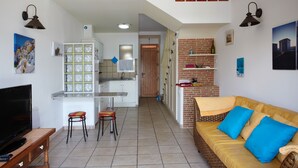 Eine moderne Küche mit weißen Schränken, ein Essbereich mit Tisch und Stühlen sowie ein Wohnzimmer mit Sofa und Kissen.