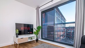 Ein modernes Wohnzimmer mit einem großen Fenster, einem Flachbildfernseher, der 'NETFLIX' anzeigt, einer kleinen Pflanze und einem grauen Teppich.