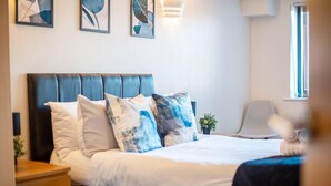 Ein ordentlich gemachtes Bett mit blauer Kopfteil, weißer Bettwäsche und Kissen mit blau-weißen Mustern. Auf dem Nachttisch steht ein kleiner Topfpflanze und in der Ecke ein Stuhl.