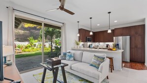 Una cocina moderna con una isla grande, un área de comedor con sofá blanco y vista a un jardín tropical.