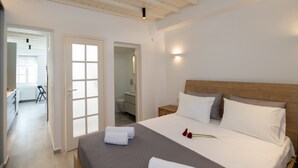 Quarto com uma cama grande, cabeceira de madeira e mesa de cabeceira. Há uma porta que dá para um banheiro e um corredor.