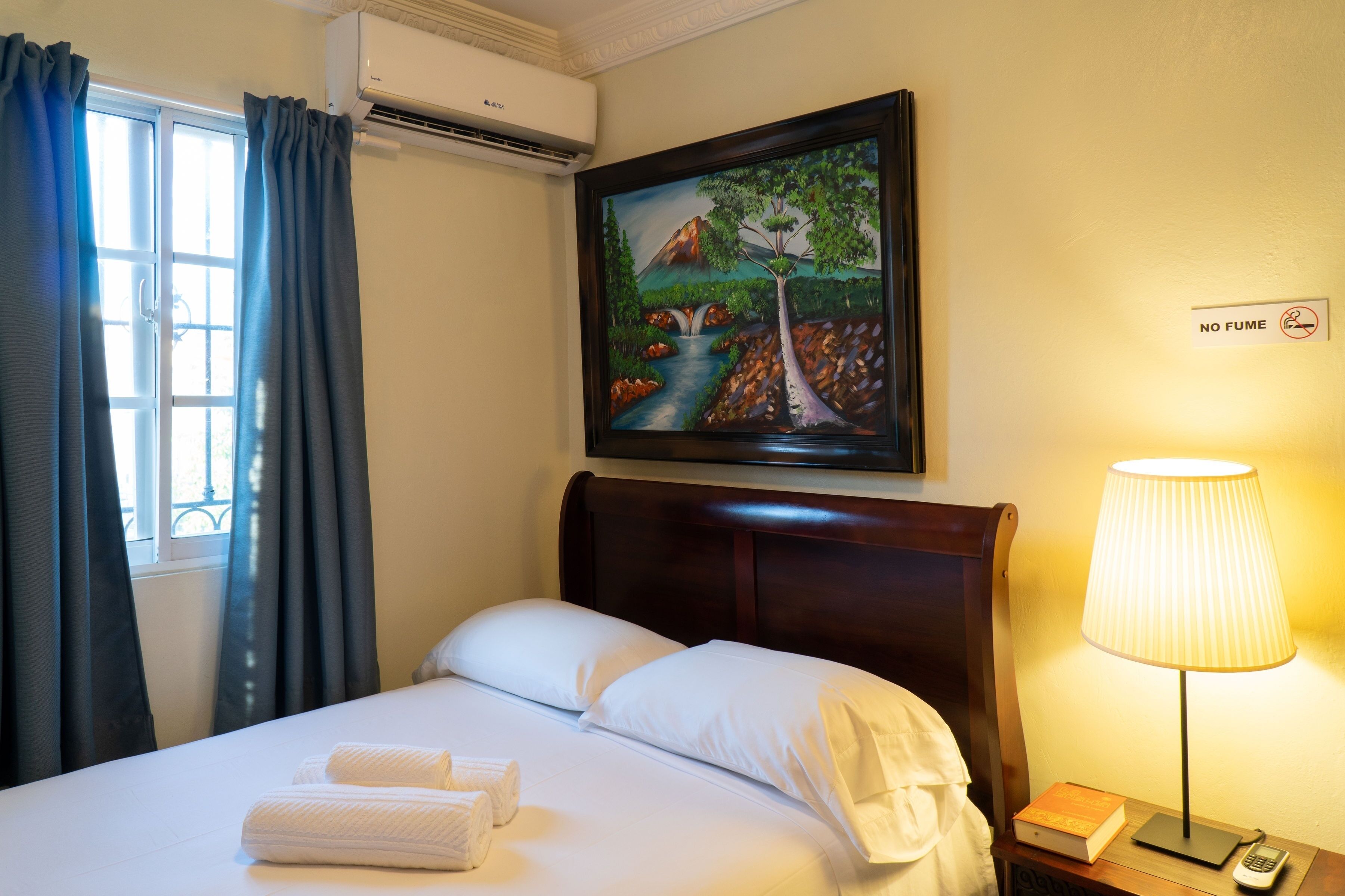 Quarto de hotel com uma cama, um quadro de paisagem, um aviso de "NÃO FUME" e uma luminária em uma mesa de cabeceira.