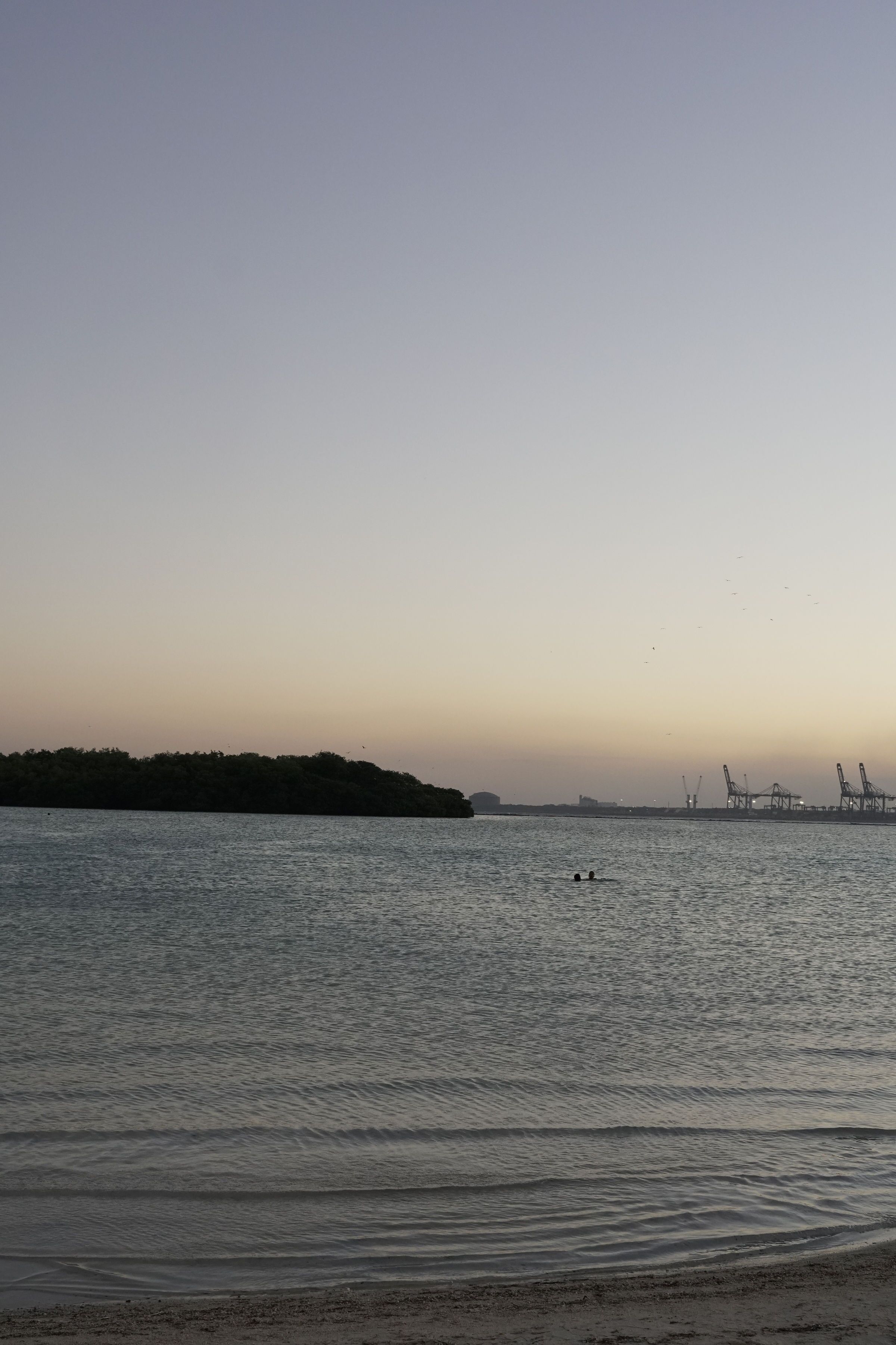 Una orilla tranquila con una costa lejana y grúas industriales.