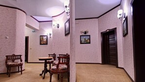 Una habitación con piso alfombrado, una mesa redonda con sillas y un banco. Hay lámparas fijadas a la pared y cuadros enmarcados.