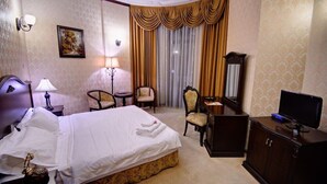 Habitación de hotel con cama, dos sillas, escritorio, televisor y un ventanal con cortinas.