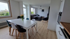 Ett modernt kök med vita skåp, en matplats med ett vitt bord och stolar samt ett vardagsrum med en soffa och ett soffbord.