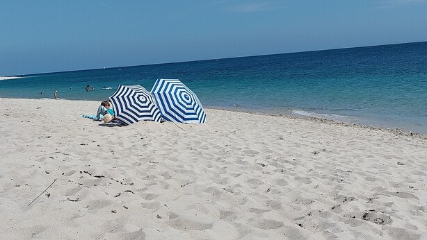 En strand med två randiga parasoller och en person som sitter på sanden.