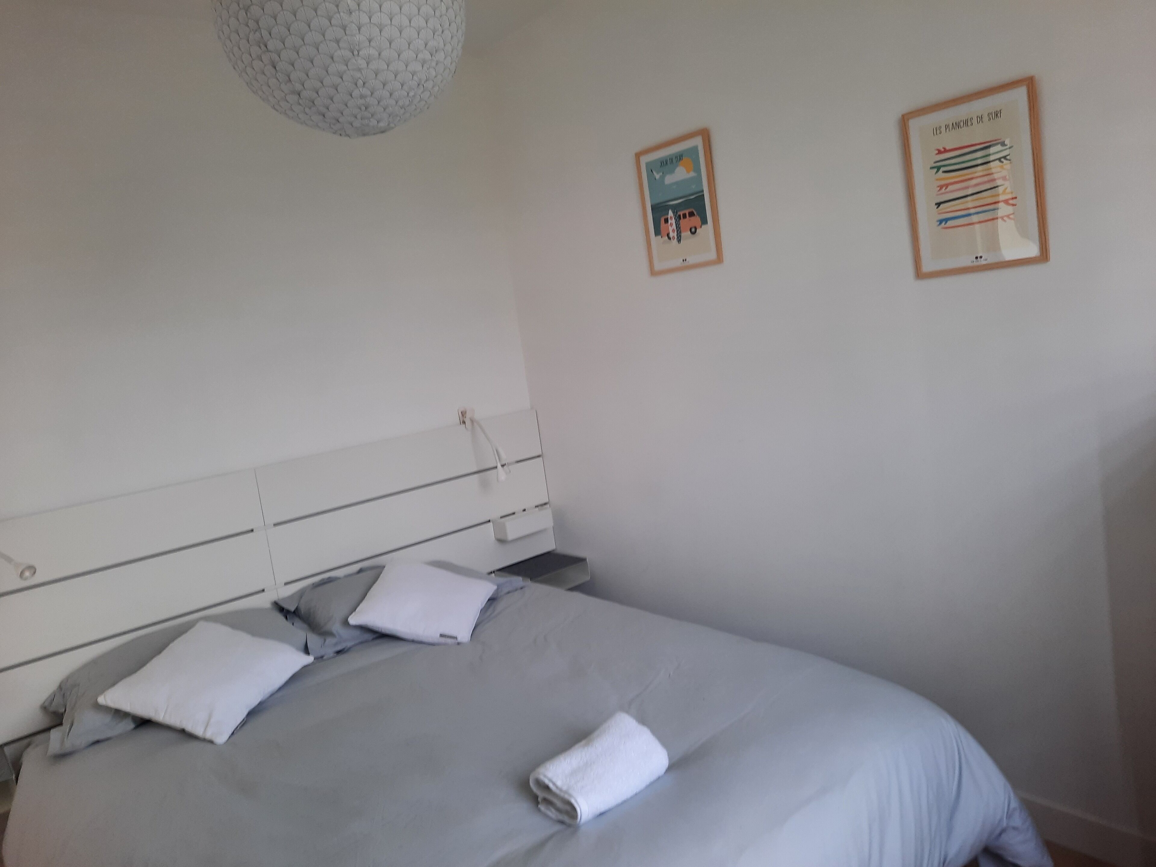 A neatly made bed with a grey duvet and white pillows, a white headboard, and a pendant light. There are two framed pictures on the wall.