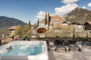 Un jacuzzi en una terraza con sillas de descanso, vista a la montaña y un edificio histórico con una torre.