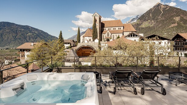 Un bain à remous sur une terrasse, avec des chaises longues, une vue sur les montagnes et un bâtiment historique doté d’une tour.