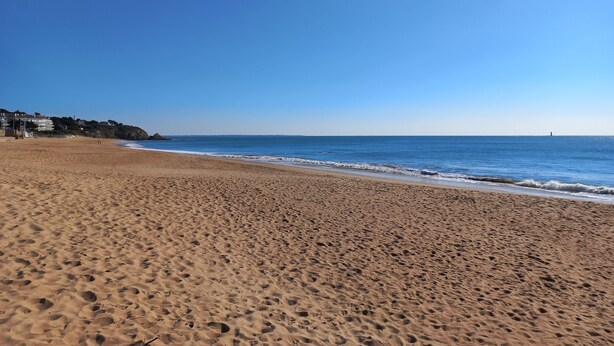 Una extensa playa de arena, cielo azul despejado, mar tranquilo y un faro a lo lejos.