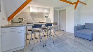 Una cocina moderna con una isla central, armarios blancos y un microondas. Hay cuatro sillas plegables y un sofá gris en la sala de estar.