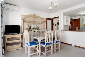 A dining area with a glass-top table, white chairs with blue cushions, a television on a wooden stand, and a decorative fireplace.