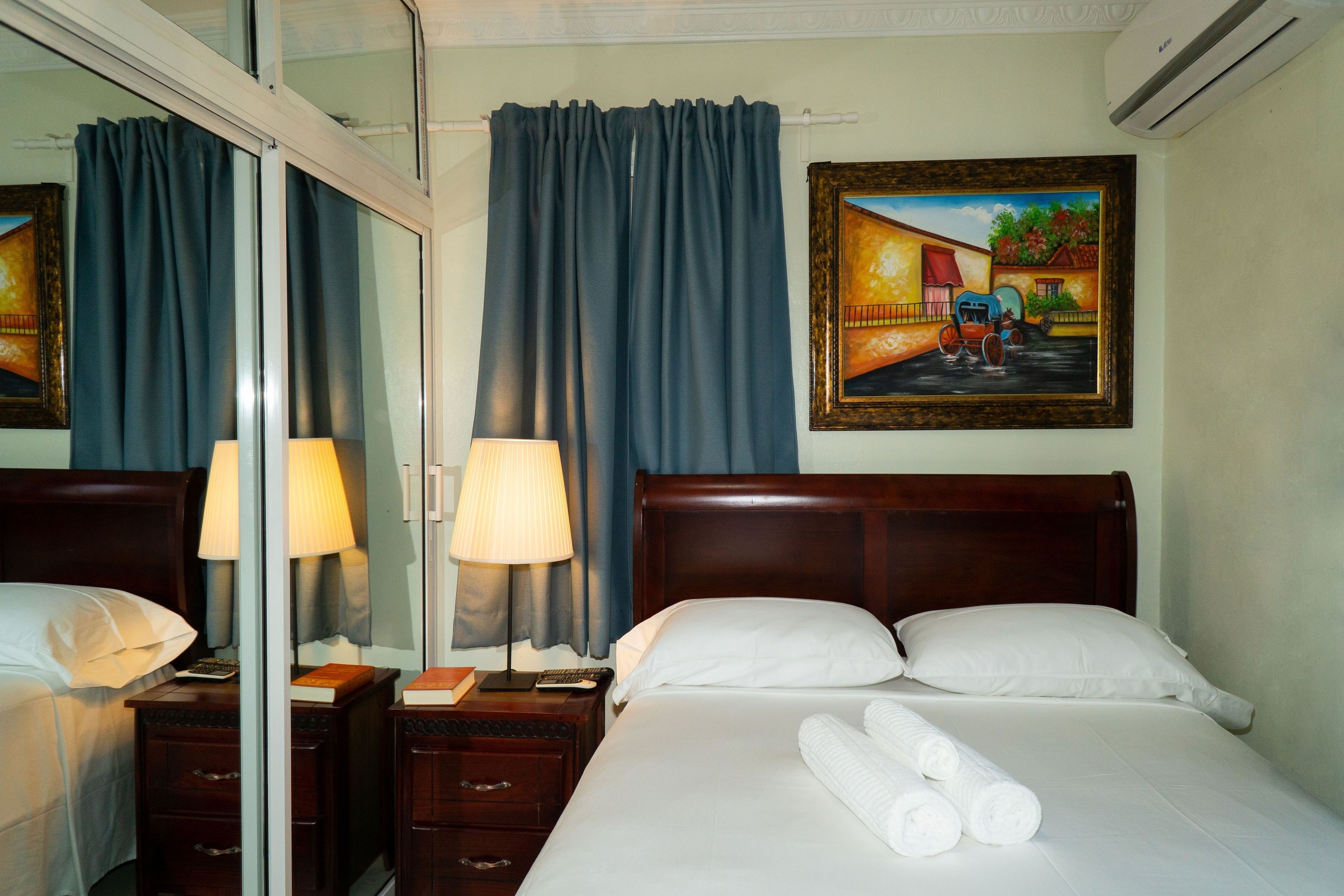 Habitación de hotel con cama, dos mesitas de noche, lámpara, un cuadro y ventana con cortinas.