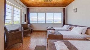 A hotel room with wooden flooring, a large window with a view, two beds, a desk, and wicker chairs. - Saint Peter Bay – 1st Floor Holiday Home with Shared Pool, Wi-Fi & Air Conditioning (Lefkimmi)