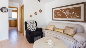 Un salon avec un canapé, un fauteuil et une table basse, une cuisine à l’arrière-plan et un grand tableau abstrait accroché au mur.