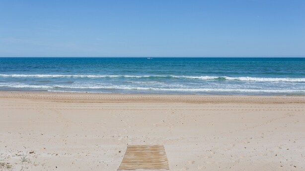 Une plage avec une promenade en bois qui mène à l’eau, un ciel dégagé et un bateau au loin.