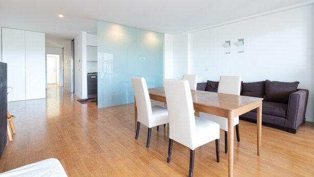 Una sala moderna con una mesa de comedor de madera, sillas blancas, un sofá oscuro y una cocina al fondo.