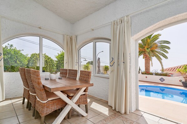 A dining area with wicker chairs and a wooden table, large arched windows, and a view of a swimming pool.