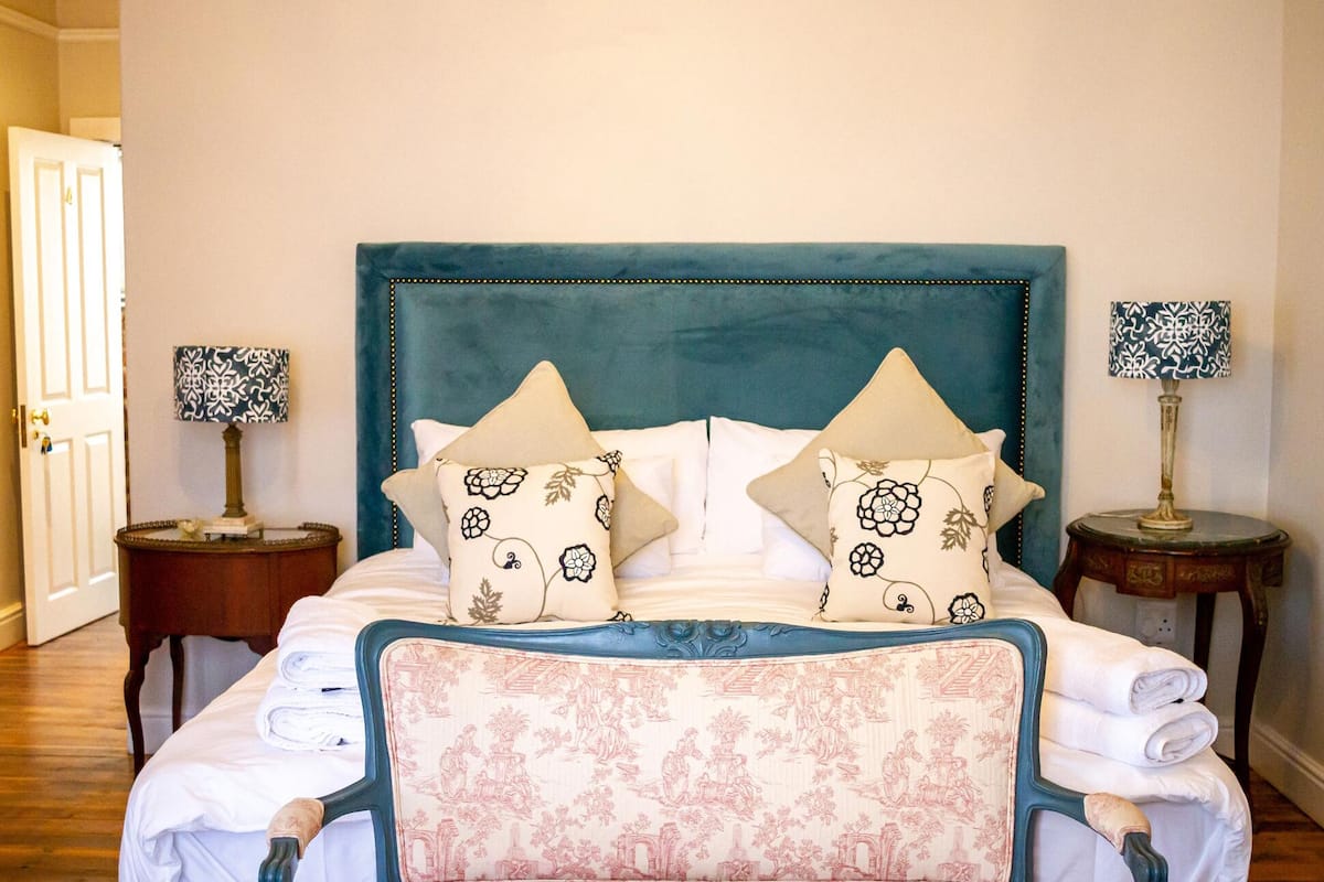 A neatly made bed with a teal headboard, two decorative pillows, and a floral-patterned chair.