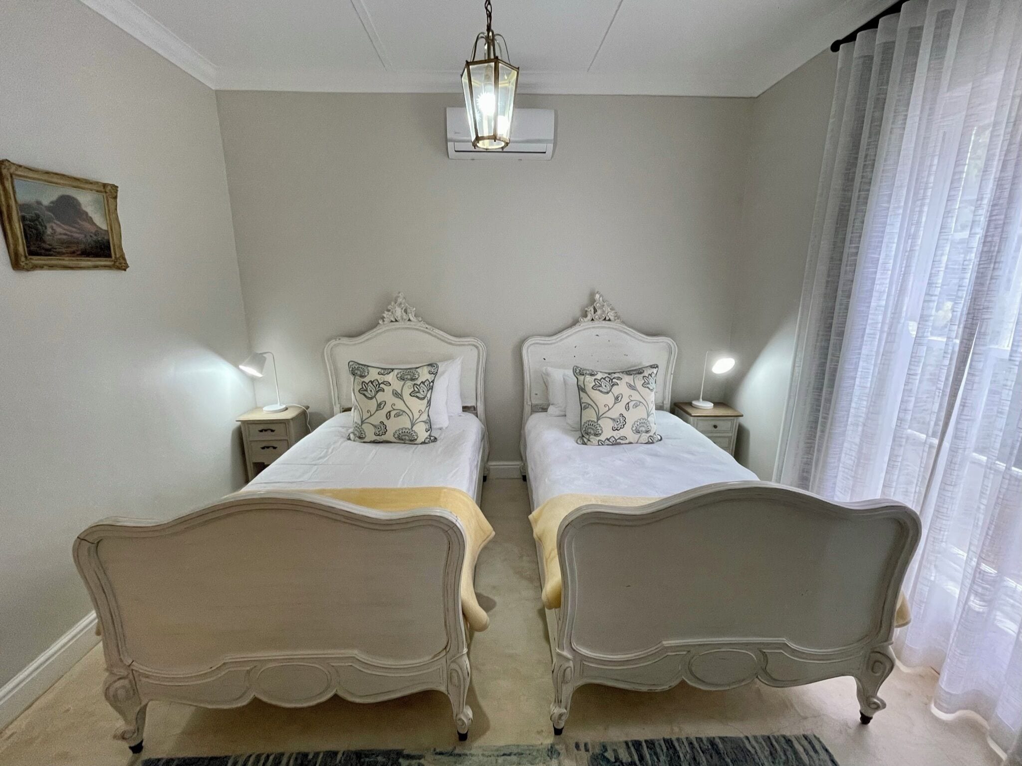 A bedroom with two beds, a hanging light fixture, a bedside table with a lamp, a framed picture on the wall, and a window with sheer curtains.