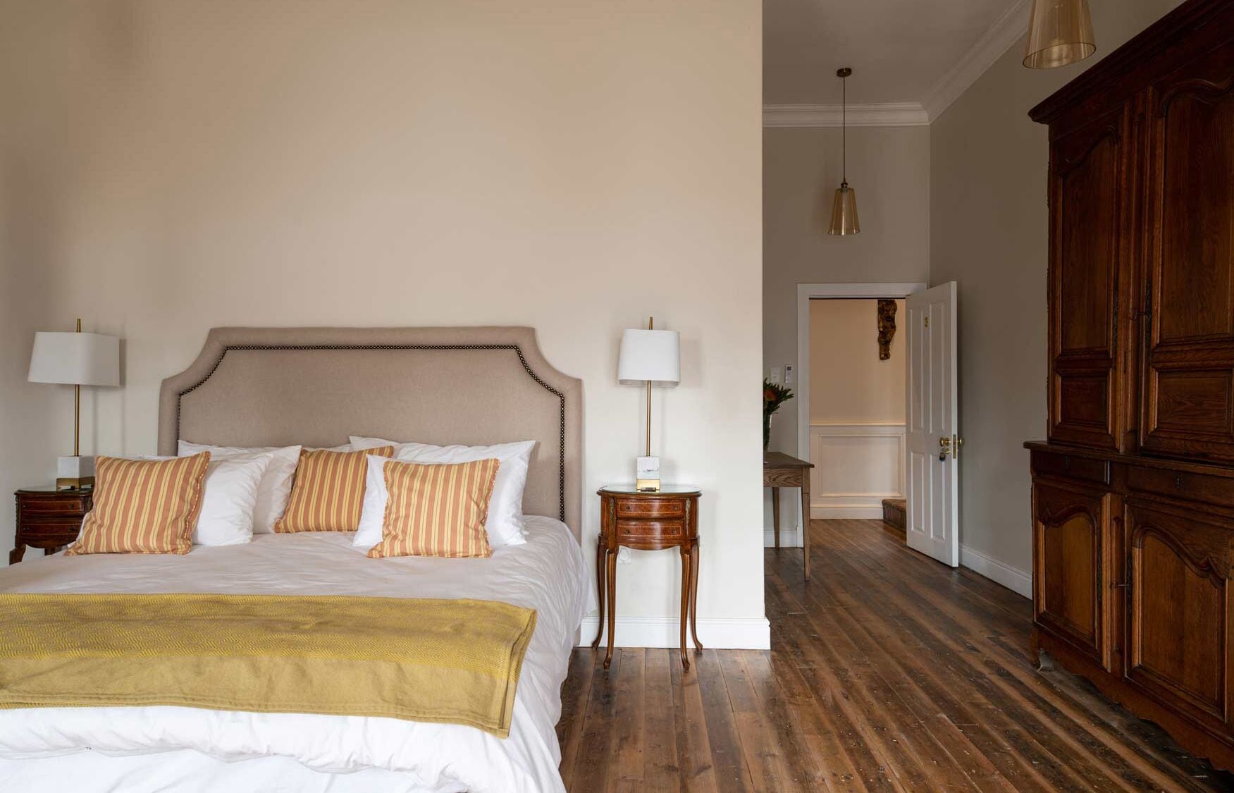 A bedroom with a large bed, two bedside tables, a wooden wardrobe, and a hallway leading to another room.