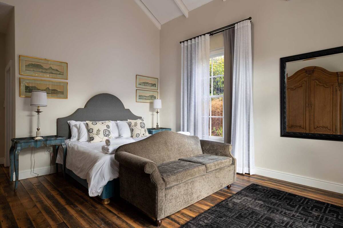 A bedroom with a bed, a sofa, a nightstand, a lamp, a window with curtains, and framed pictures on the wall.