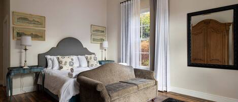 A bedroom with a bed, a sofa, a nightstand, a lamp, a window with curtains, and framed pictures on the wall.
