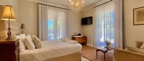 A well-lit bedroom with a large bed, a bedside table with lamps, a TV, and a view of greenery outside.